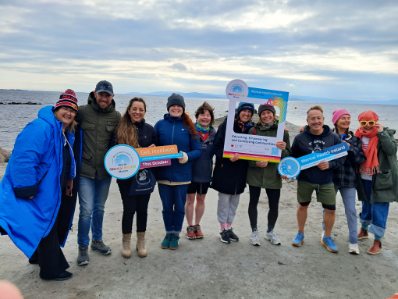 group of people wrapped up in warm clothes with Mental Health Ireland merchandise in their hand standing on a beach