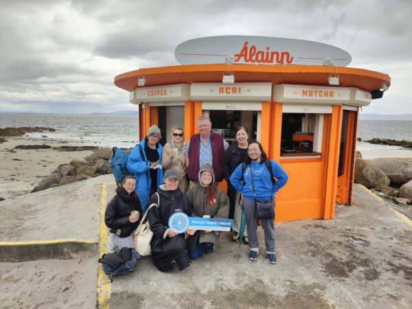 A group of people standing outside a hut on the beach holding Mental Health Ireland hand held signs